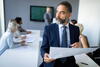 a man wearing a suit and tie holding a large piece of paper with coworkers sitting at a conference table behind him